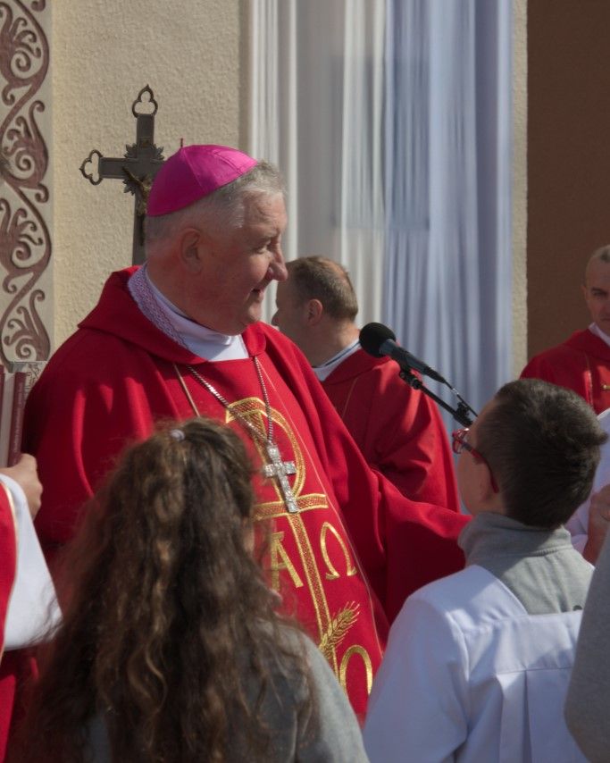 Biskup Romuald Kamiński w czerwonym ornacie przemawia do ludzi podczas uroczystości religijnej na zewnątrz kościoła.