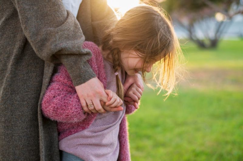 Mała dziewczynka w różowym swetrze i w paskowanym topie przytula się do nogi dorosłego w szarym płaszczu na tle zielonego parku w promieniach zachodzącego słońca.